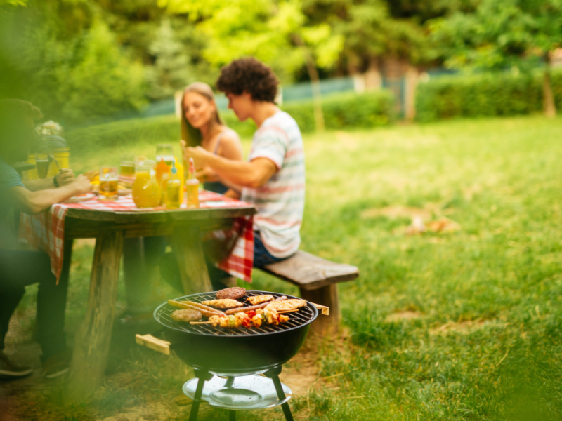 Ein paar Menschen sitzen an einem Tisch und essen. Im Vordergrund sieht man einen Grill mit Grillgut darauf.