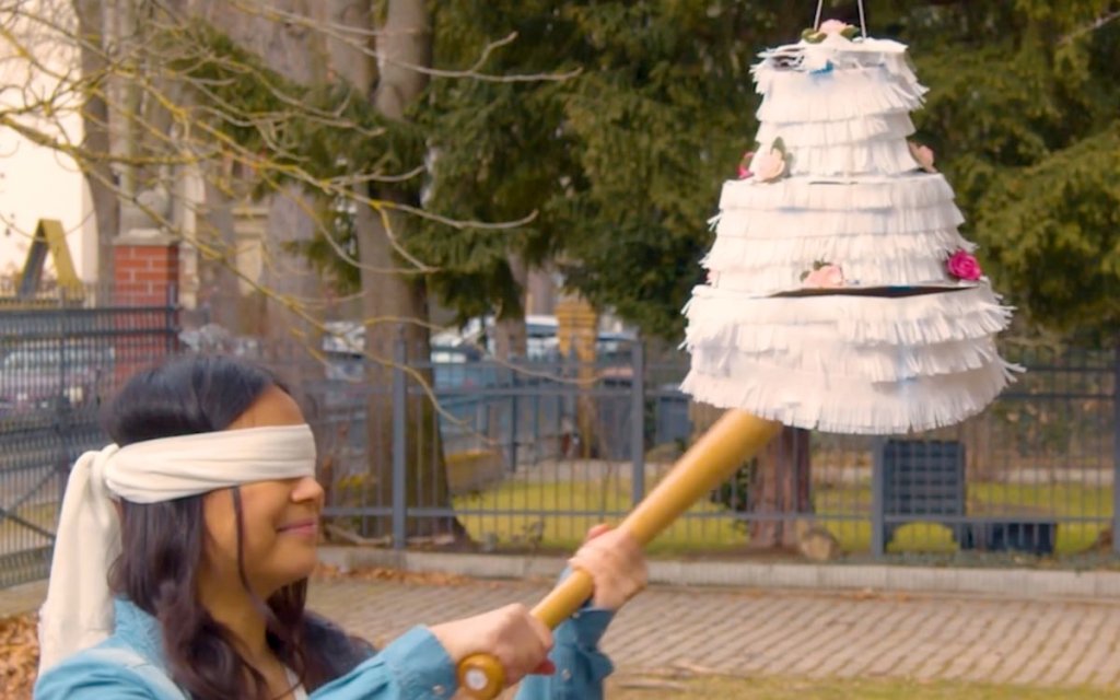 Eine junge Frau mit verbundenen Augen schlägt auf eine Piñata in Form einer Hochzeitstorte.