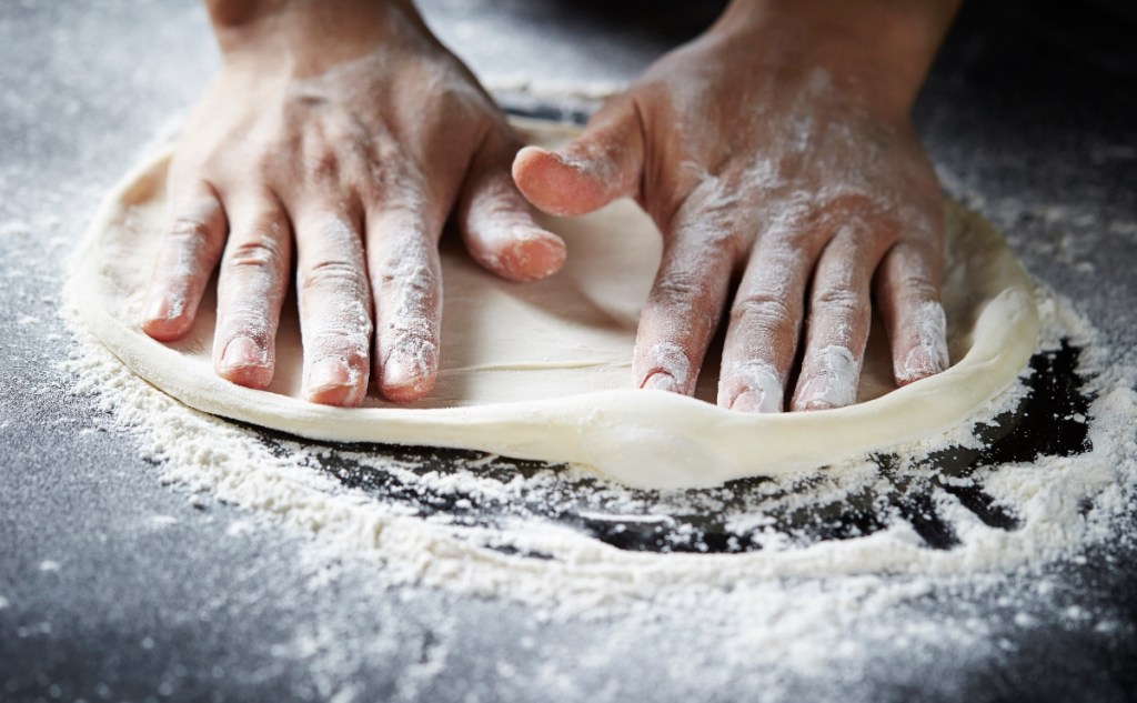 Ein Pizzateig wird auf einer mit Mehl bestäubten Unterlage geknetet.