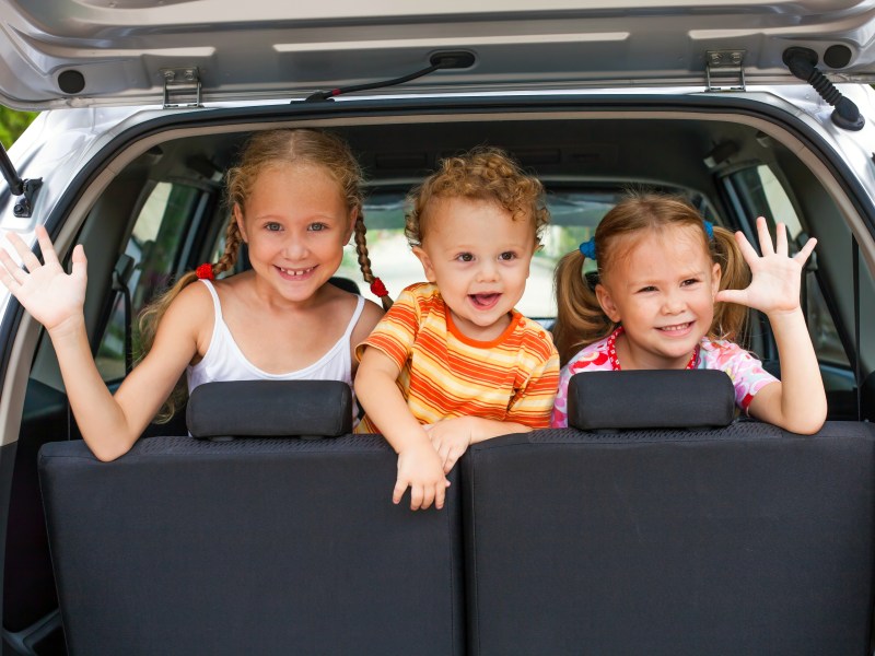 Drei kleine Kinder sitzen auf dem Rücksitz eines Autos und winken durch die geöffnete Kofferraum-Klappe.