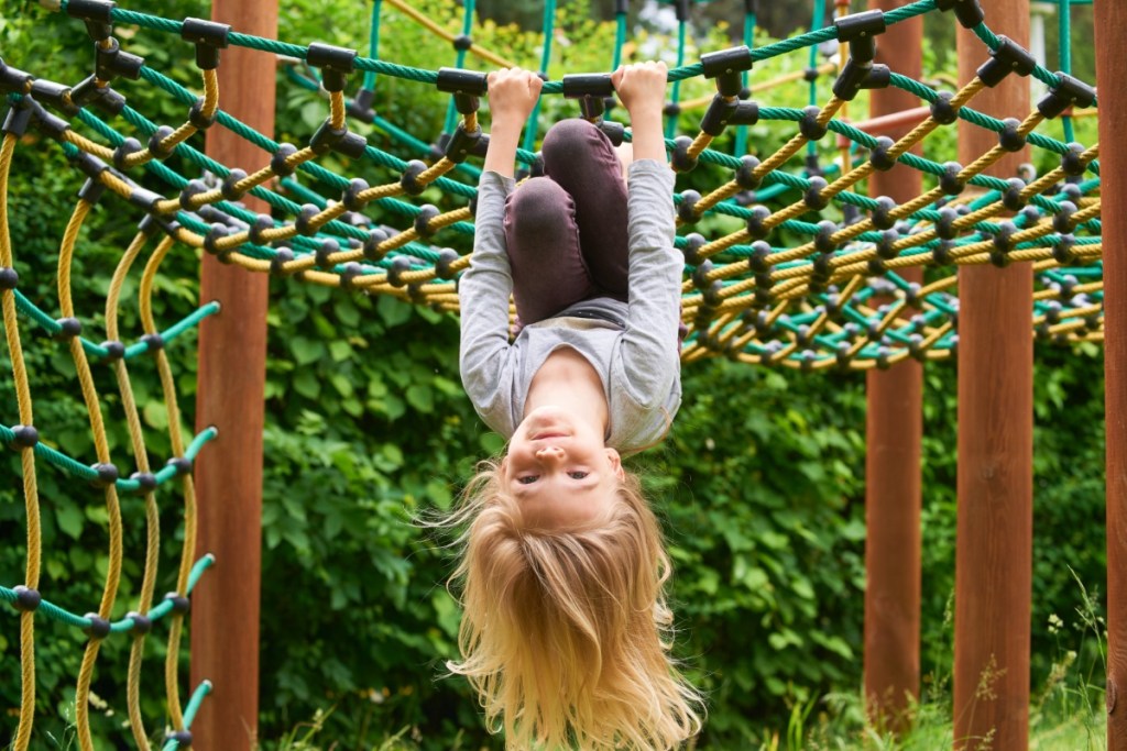 Ein kleines, blondes Mädchen spielt auf einem Spielplatz und turnt in einem Kletterseil.