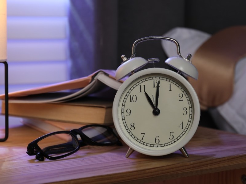 ein Wecker, eine Brille und ein Buch neben einem Bett