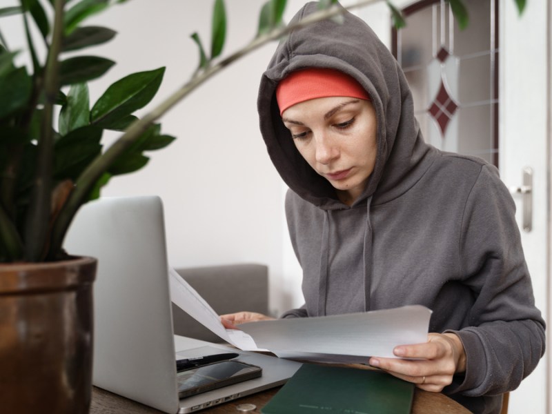 Eine Frau sitzt am Schreibtisch und schaut sich Papiere an. Sie trägt eine Mütze und darüber hat sie die Kapuze ihres Hoodies gezogen.