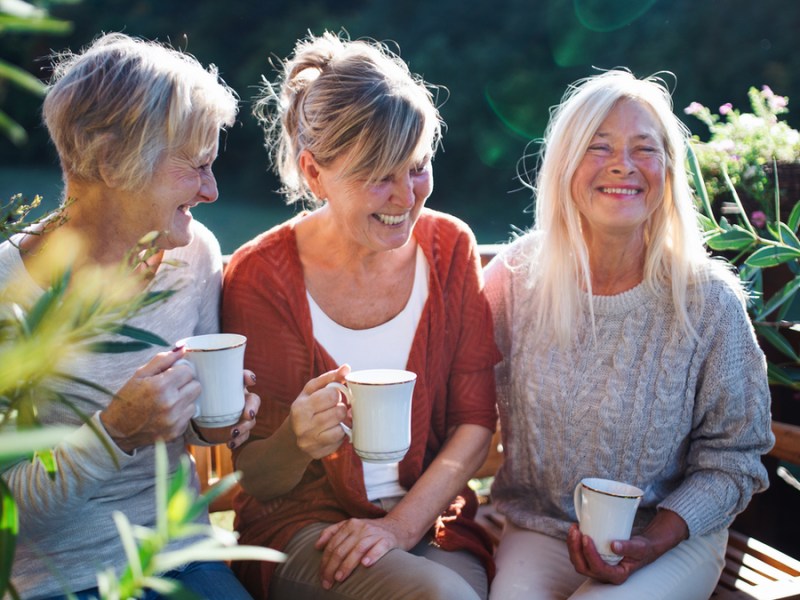 Drei Frauen mittleren Alters sitzen draußen auf einer Bank und lachen.