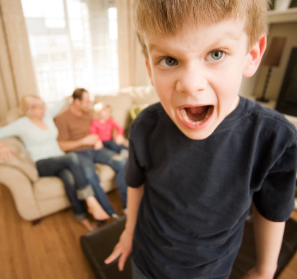 Kleiner Junge schreit in die Kamera, während hinter ihm seine Familie auf dem Sofa sitzt