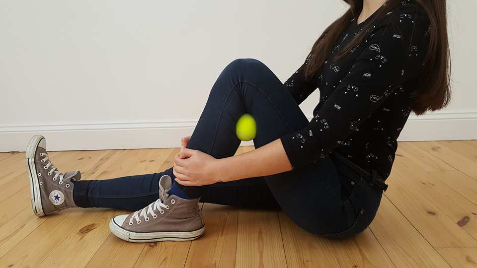 Eine junge Frau klemmt einen Tennisball zwischen die Kniekehle.