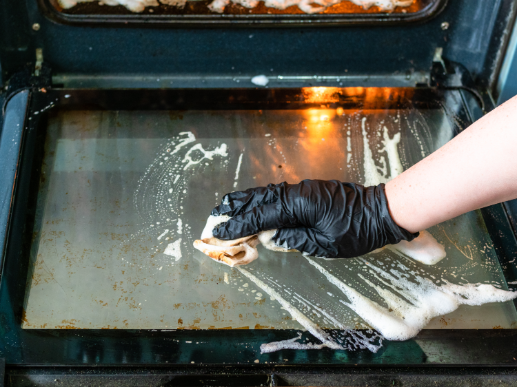 Hand mit Putzhandschuh reinigt eingebrannte Fettspritzer am Backofen.