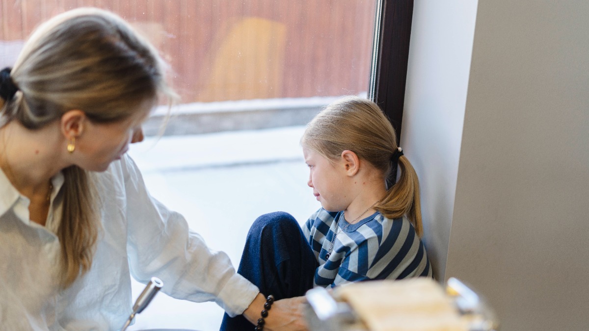 Mutter spricht auf Tochter ein, die traurig aus dem Fenster schaut