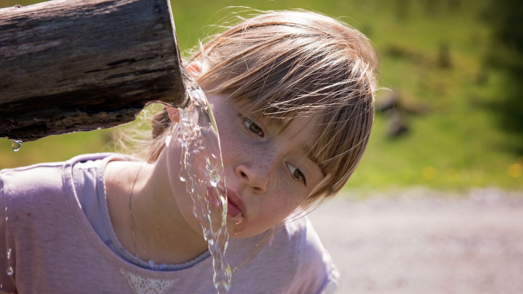 Ein Mädchen trinkt Wasser aus einer hölzernen Wasserleitung.