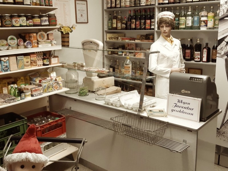 Eine Frau steht hinter der Theke eines Konsums. Das Bild ist in Sepia gehalten.