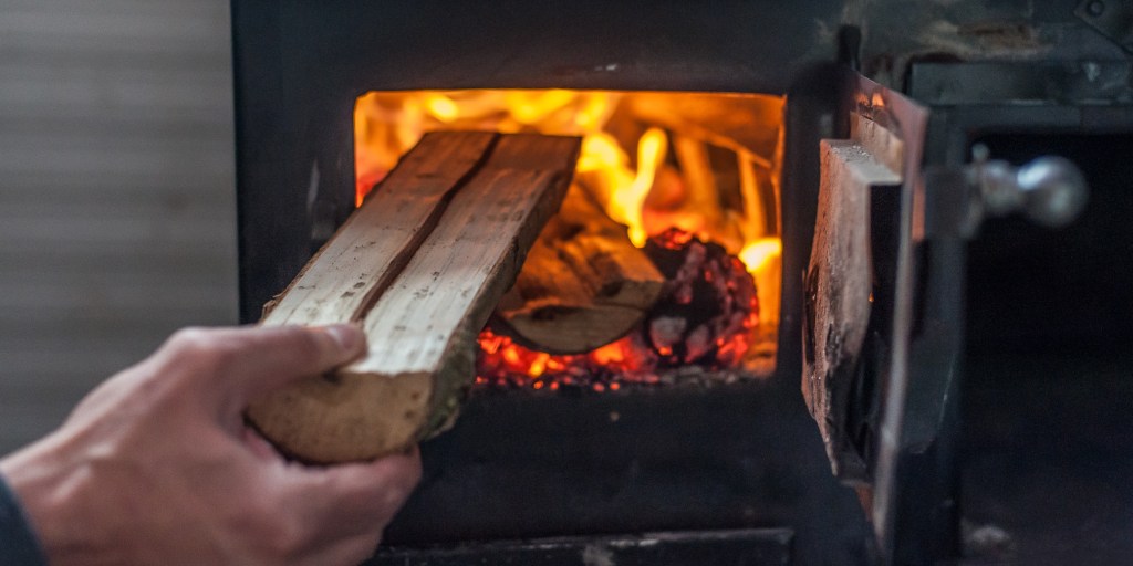 Jemand legt ein Holzscheit in einen brennenden Kaminofen.