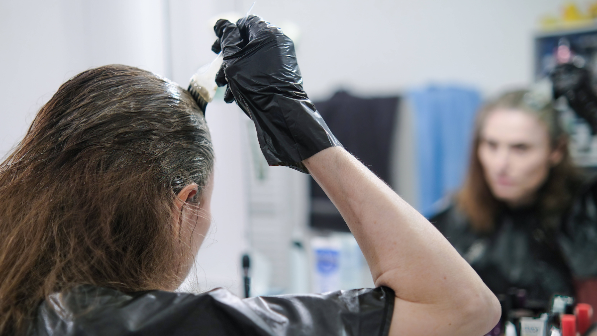 Eine Frau steht vor einem Spiegel und trÃ¤gt Haarfarbe mit einem Pinsel auf.