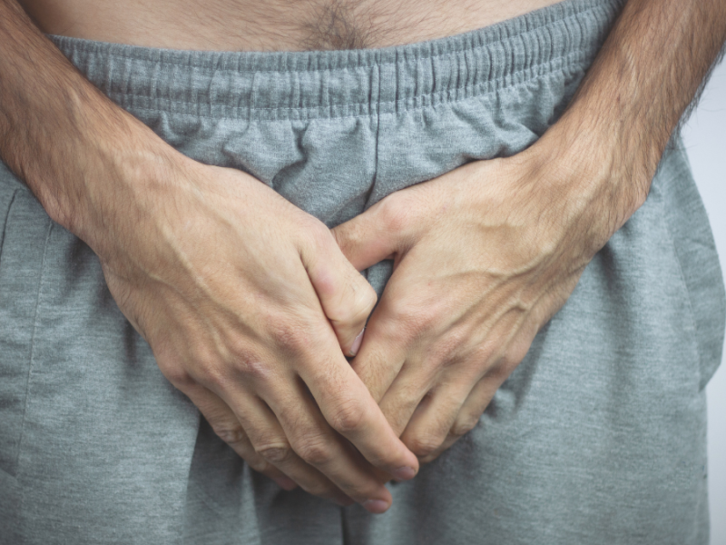 Ein Mann hält auf Höhe seines Geschlechtsteils beide Hände an seine graue Jogginghose.