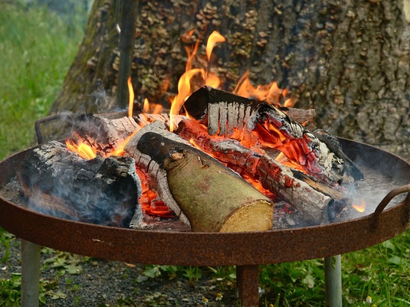 Eine Feuerschale, in der ein Feuer brennt, steht in einem Garten vor einem Baum.