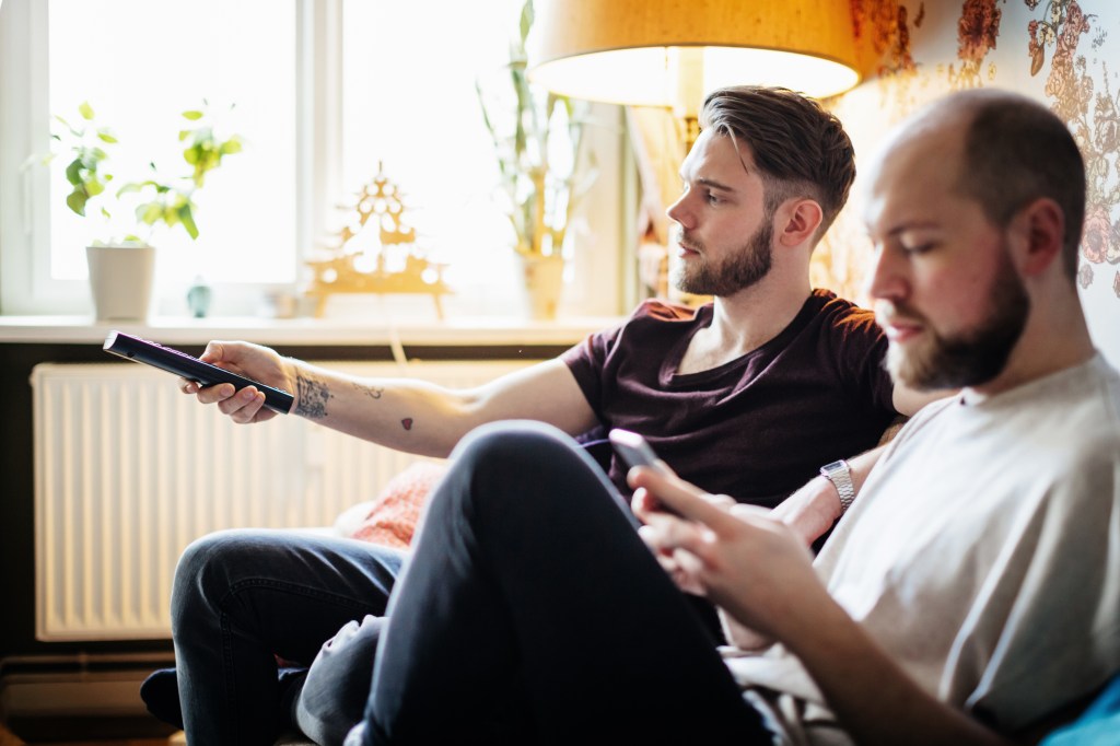 Zwei junge Männer sitzen auf einem Sofa, einer hält Fernbedienung eines Fernsehers.