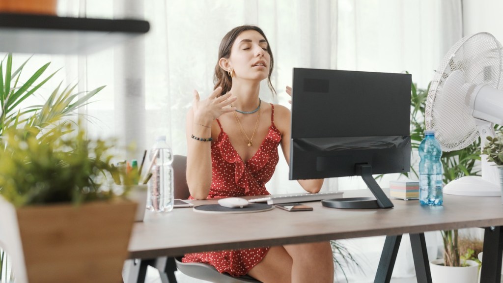 Frau mit luftigem Outfit in einem Büro vor einem Ventilator am Schreibtisch.