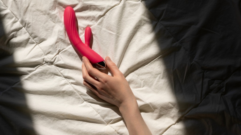 Eine Hand greift nach einem roten Vibrator, der auf dem Bett liegt.