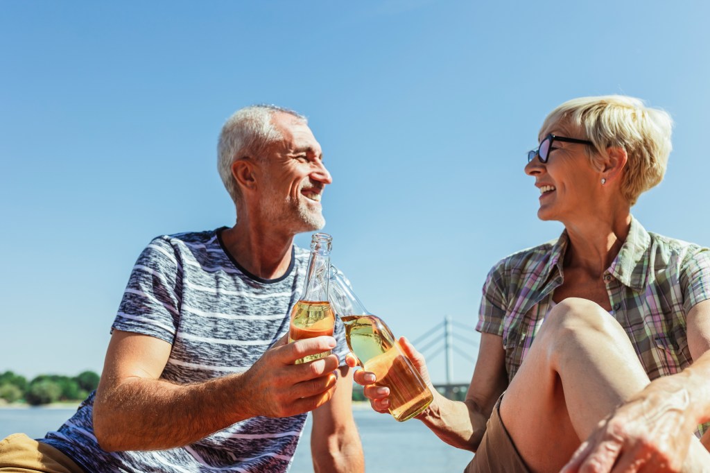 Ein älteres Paar stößt an einem Fluss mit Bier an.