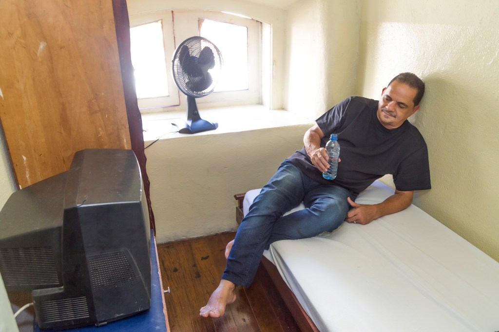 Mann sitzt bei Hitze auf einem Bett vor einem Ventilator.