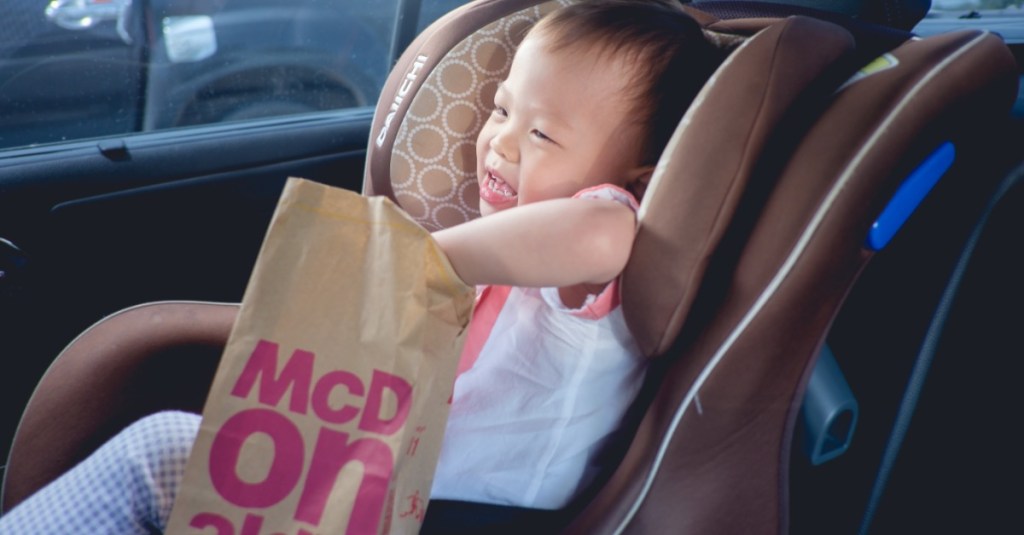 kleines Kind sitzt im Kindersitz im Auto und freut sich über eine McDonalds Tüte