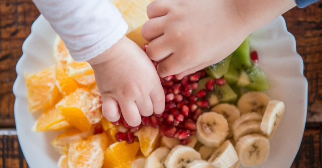 Snackteller mit frischem Obst und zwei Kinderhände, die etwas davon wegnehmen