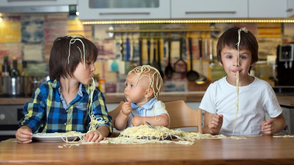 drei Kinder sitzen am Tisch und haben Spaghetti auf dem Kopf