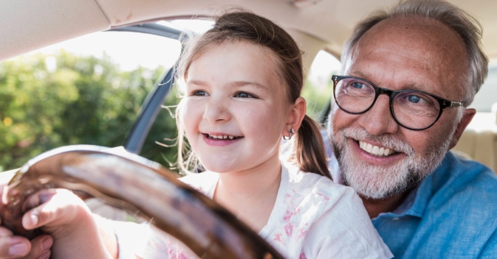 kleines Mädchen sitzt im Auto auf Opas Schoß, während sie das Lenkrad festhält