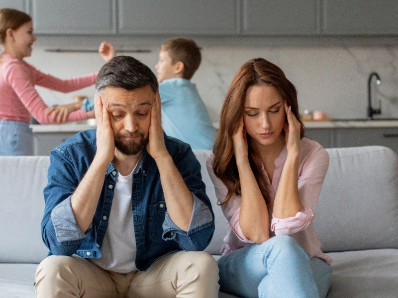 Eltern sitzen genervt auf der Couch, wÃ¤hrend sich die Kinder im Hintergrund streiten