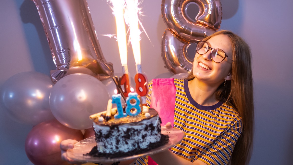 ein MÃ¤dchen hÃ¤lt eine Torte in der Hand, im Hintergrund sind zwei Luftballons zu sehen, die eine 18 zeigen