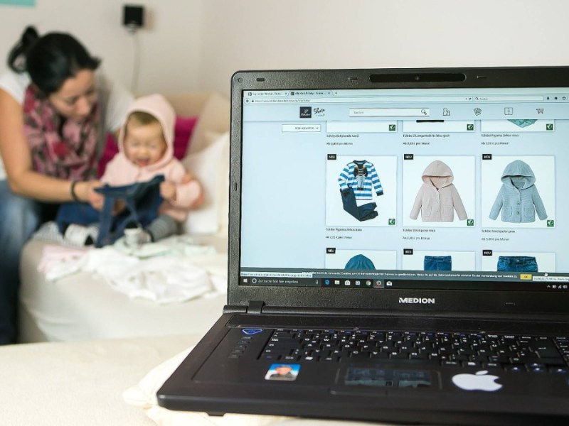 Ein Laptop mit einer Seite fÃ¼r Kindermode ist zu sehen, dahinter sitzt eine Mutter mit ihrem Baby auf dem Sofa