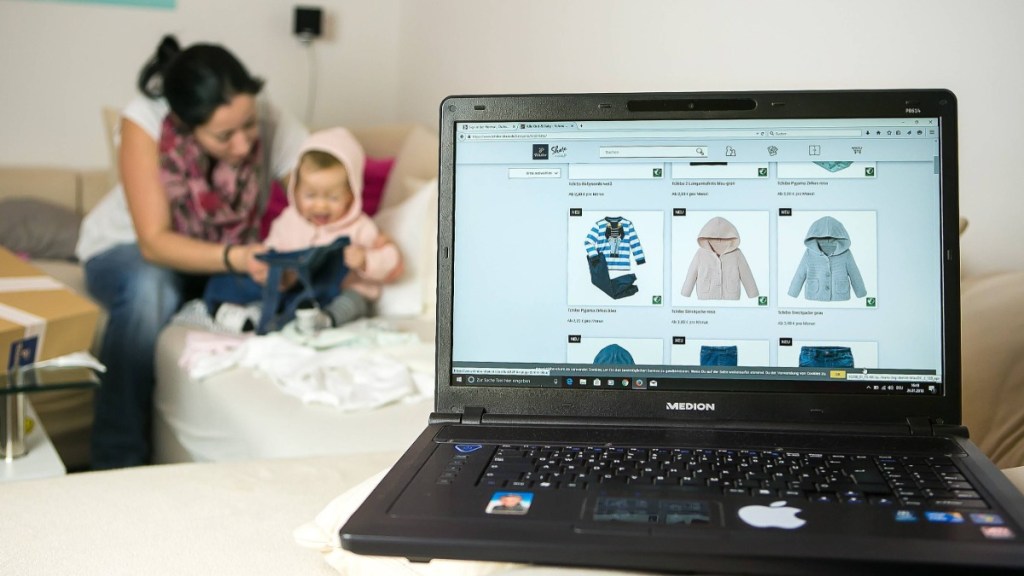 Ein Laptop mit einer Seite für Kindermode ist zu sehen, dahinter sitzt eine Mutter mit ihrem Baby auf dem Sofa