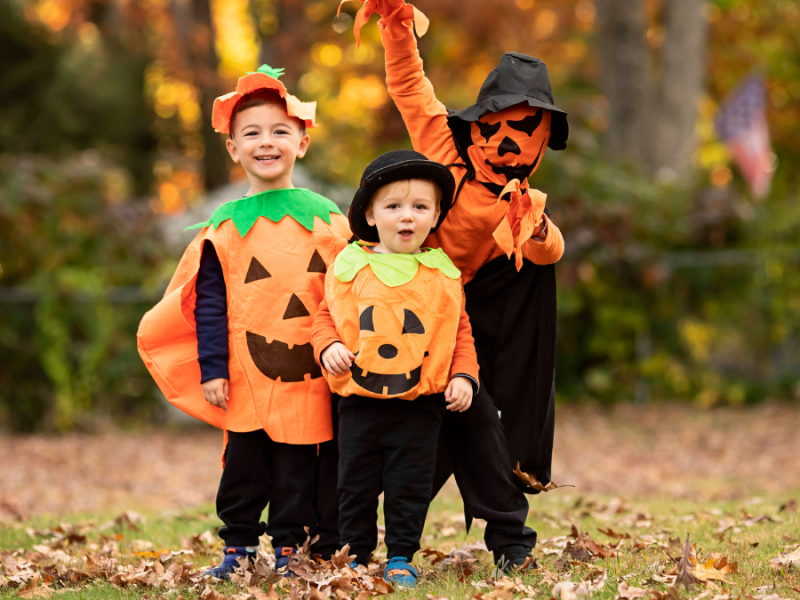 Drei Kinder mit KÃ¼rbiskostÃ¼men auf einer Wiese mit buntem Herbstlaub