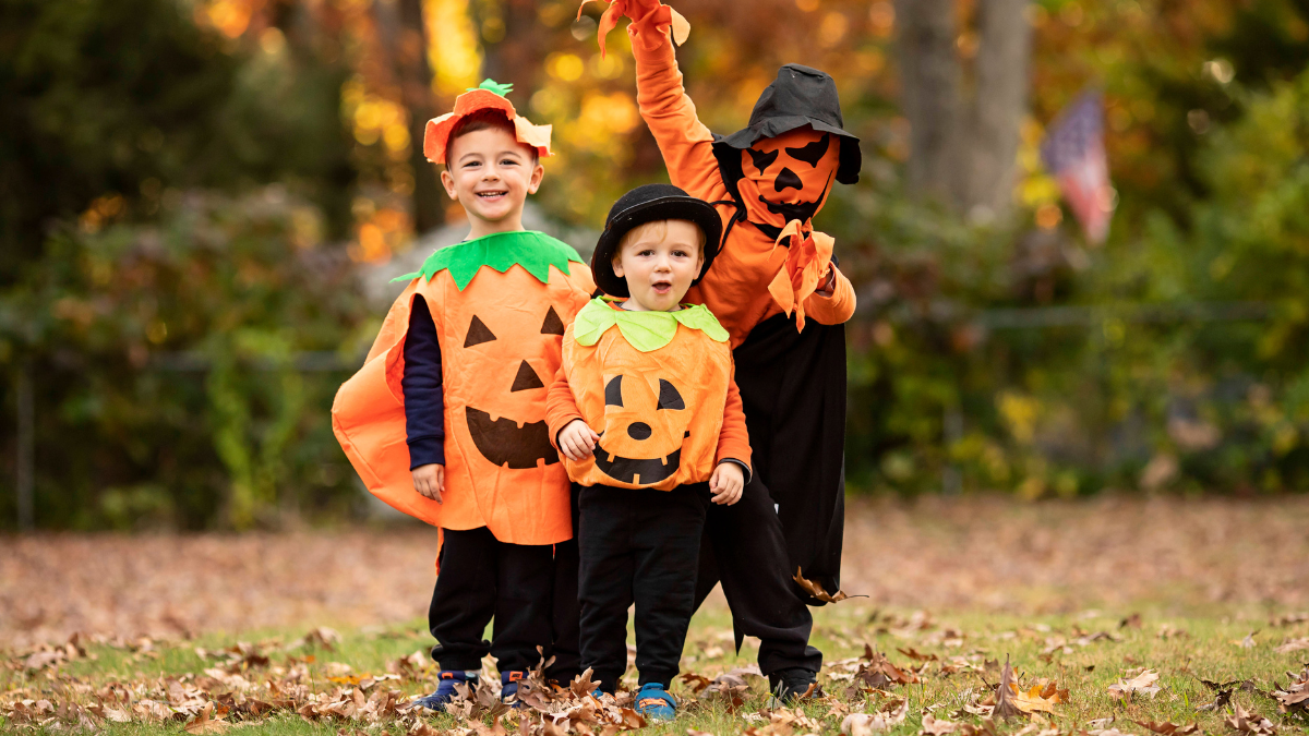 Drei Kinder mit KÃ¼rbiskostÃ¼men auf einer Wiese mit buntem Herbstlaub