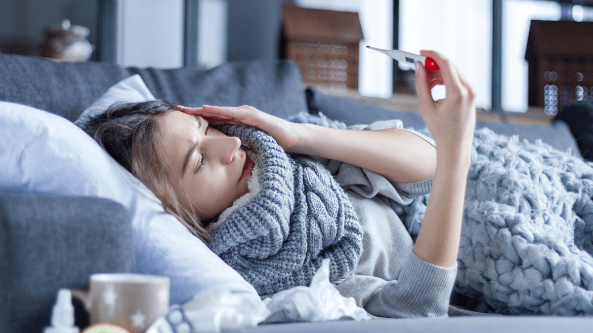 Eine Frau liegt krank auf dem Sofa und schaut auf ein Fieberthermometer.