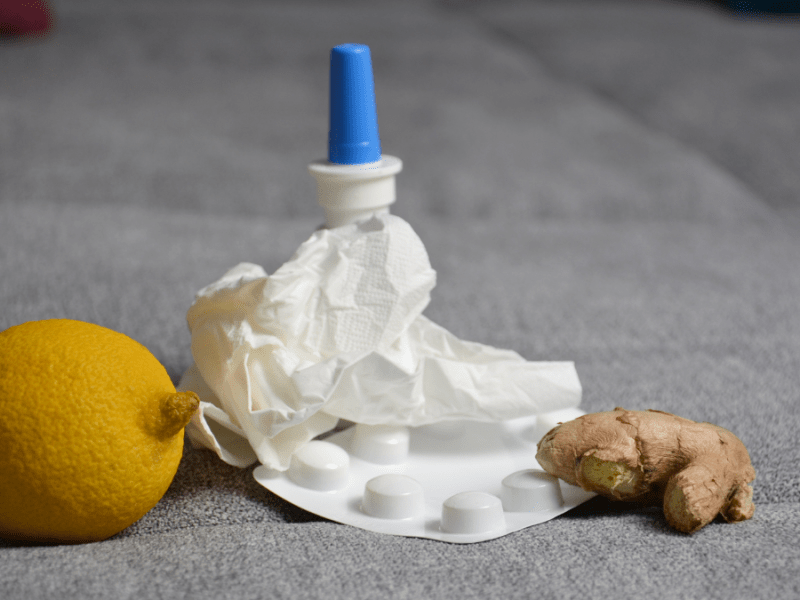 Eine Zitrone, ein Nasenspray, ein Medikamentenblister, ein zerknülltes Taschentuch und ein Stück Ingwer liegen auf einer grauen Couch.