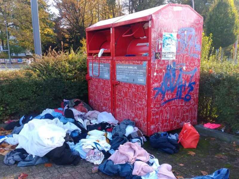 Ein roter Altkleidercontainer, vor dem Kleidung verstreut liegt.