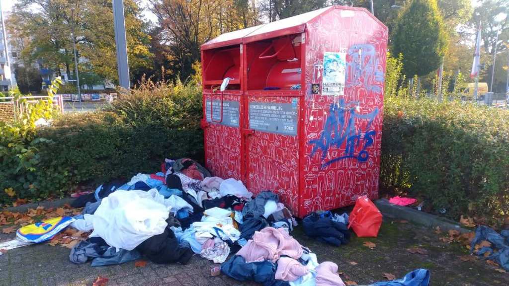 Ein roter Altkleidercontainer, vor dem Kleidung verstreut liegt.