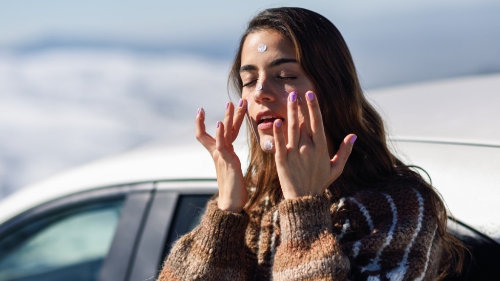 Eine Frau steht im Freien vor ihrem Auto und cremt ihr Gesicht ein.