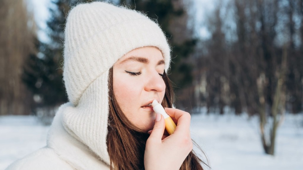 Eine Prson trägt Lippenpflege auf ihre Haut auf. Sie steht draußen und hat Wintersachen und eine Mütze auf.