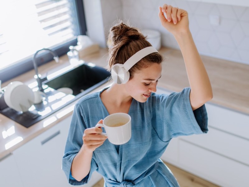 Eine Frau hat Kopfhörer auf und eine Tasse in der Hand und tanzt in ihrer Küche.