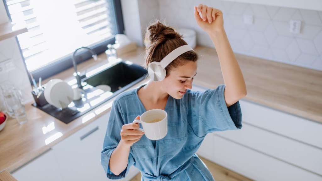 Eine Frau hat Kopfhörer auf und eine Tasse in der Hand und tanzt in ihrer Küche.