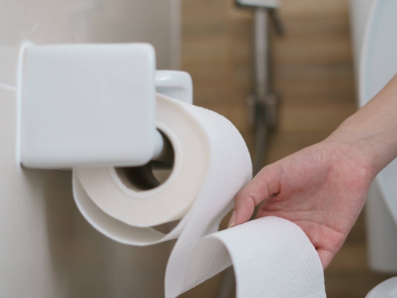Eine Hand zieht Klopapier von einer Rolle in einem Badezimmer ab.