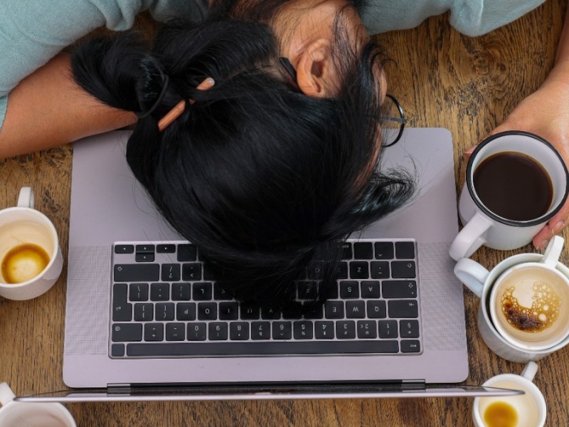 Eine Frau umgeben von leeren Tassen, die müde mit dem Kopf auf der Tastatur eines Laptops liegt.
